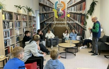 W bibliotece młodzież siedzi w okręgu, pośrodku stoi mężczyzna tłumaczący im hip hop